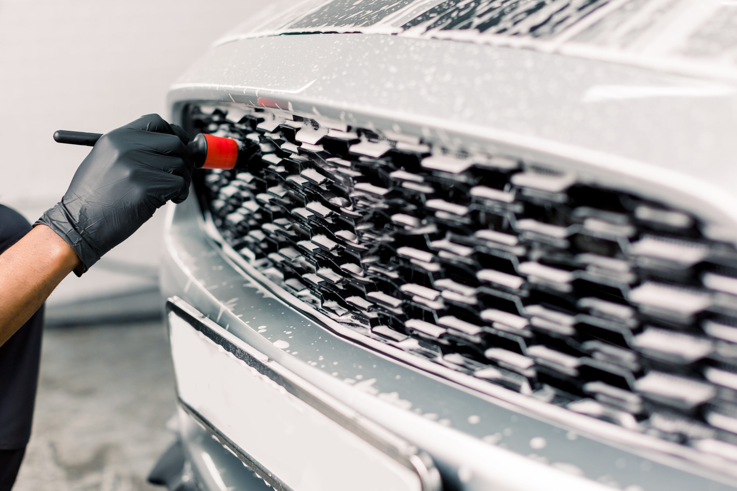 Car detailing wash. Detailing cleaning with soap at car wash. Cropped image of hand of male worker in rubber gloves washing car radiator grille of luxury white car with special brush